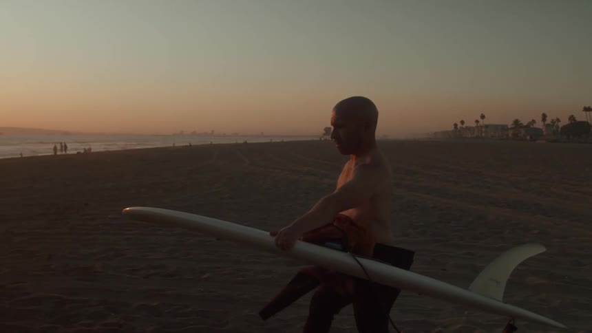 An older white man is walking on the beach as they are holding there surfboard