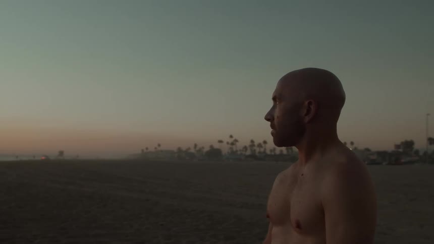 A man stands bare chested on a beach and looks out at the water