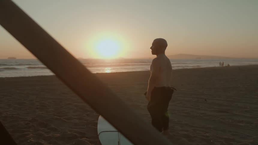A surfer looks out towards the ocean while standing next to a surfboard on a beach in the evening