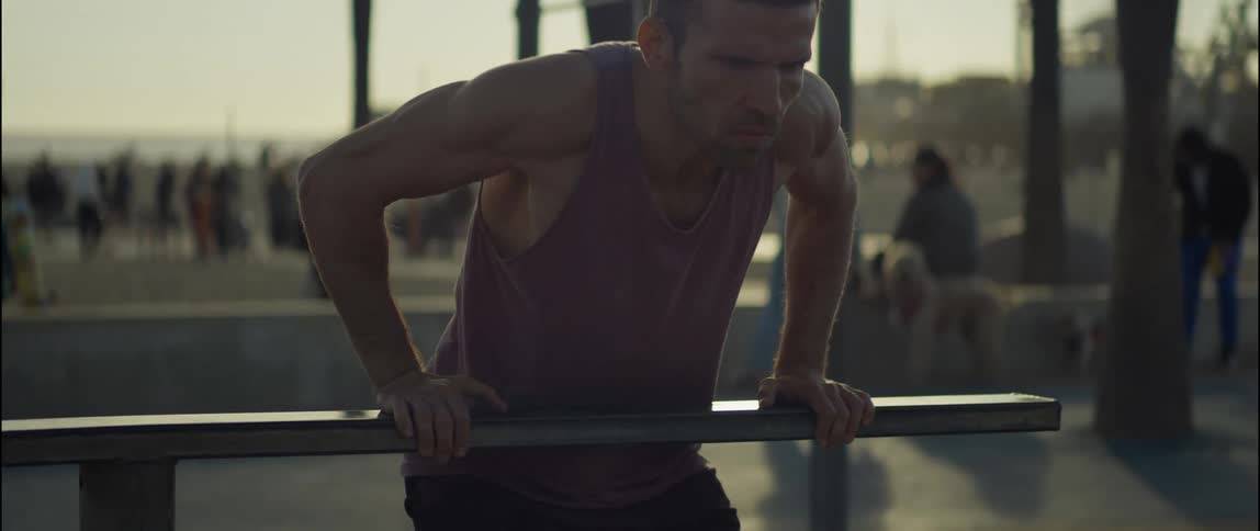 A man does push ups on a bar at the beach with a crowd of people behind him