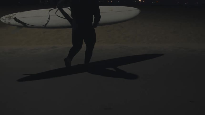 A man carries his surfboard on the beach at night