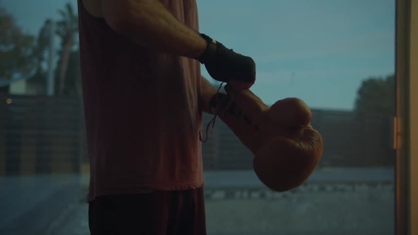 A man is lacing up boxing gloves in a room facing a yard