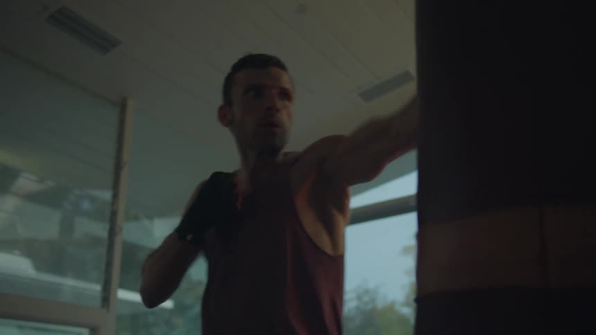 A man works out punching a hanging heavy bag at home near large windows