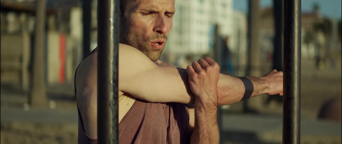 A man does arm stretches while standing before exercise equipment on a beach during the day