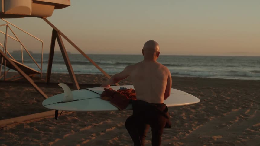A man carrying his surfboard with towel and wax on top toward the lifeguard tower and ocean waves