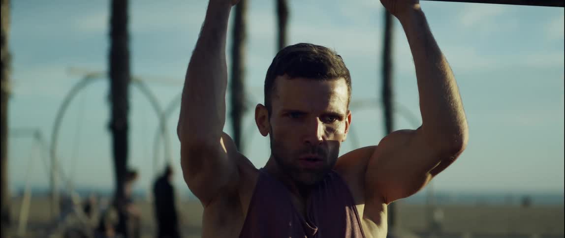 A man is working out on equipment at the beach as other people walk around behind him