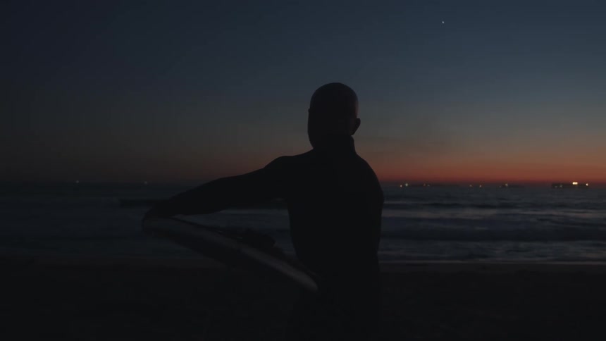 A man carries a surfboard while walking across a beach at night