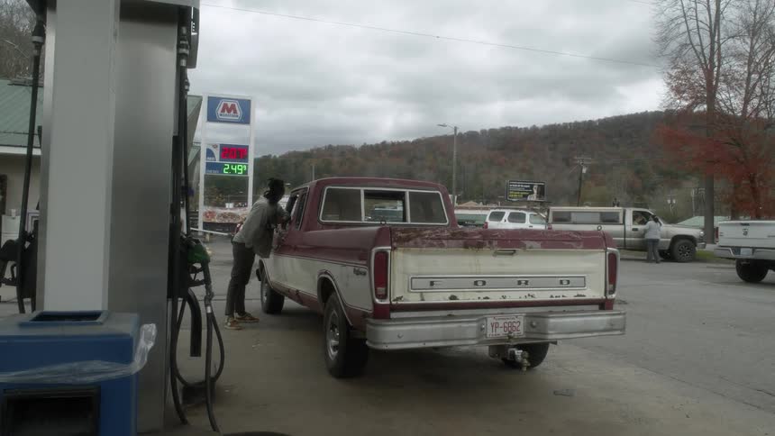 Young man talking to a person sitting on the driver seat of a pickup truck in a gas station of a small town