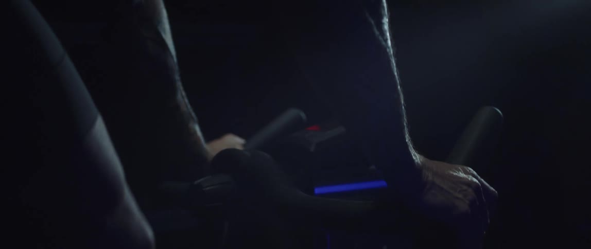 Athletic man gripping the handlebar of the stationary bike that hes riding in a dark room