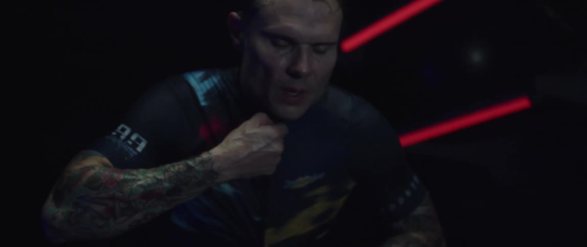 Man zips his shirt then bends forward tired in room lit by red neon light