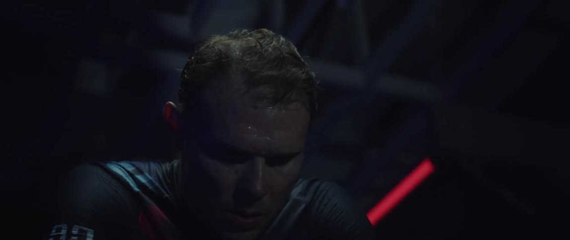 Close up of the face of a tired man pedaling a static bicycle inside a dimly lit room
