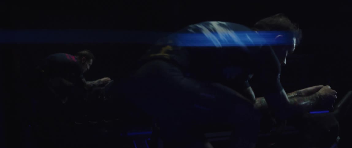 Focused man riding a stationary bike next to a mirror reflecting him in a dark gym room