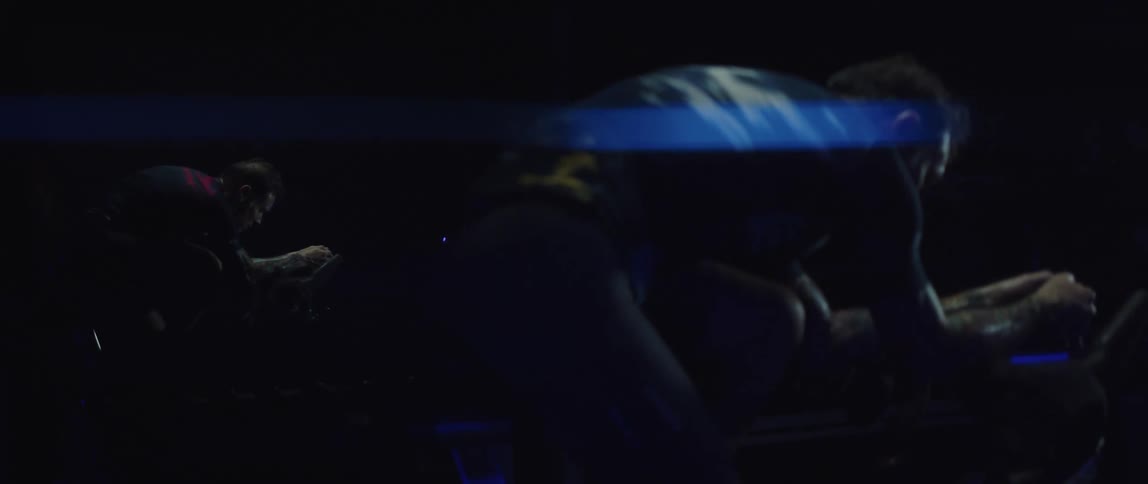 Focused man riding a stationary bike next to a mirror reflecting him in a dark gym room