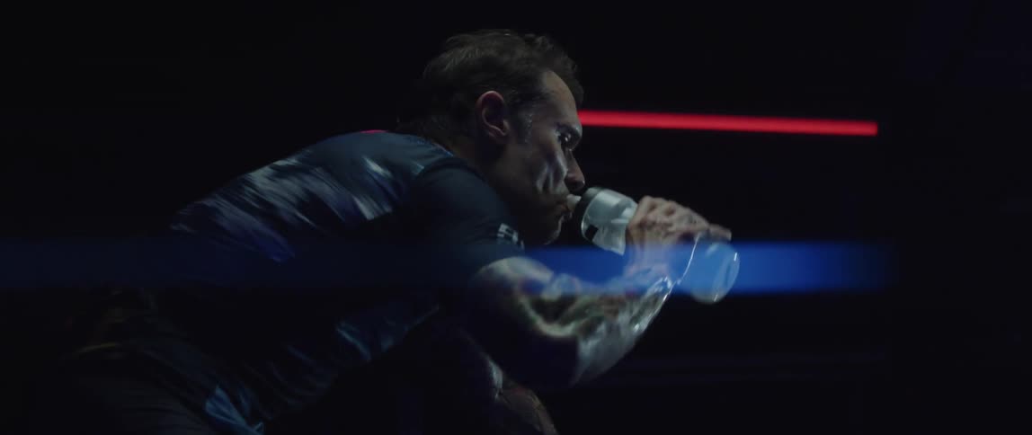 A man takes a sip from a water bottle while cycling on an exercise bike in a dark room