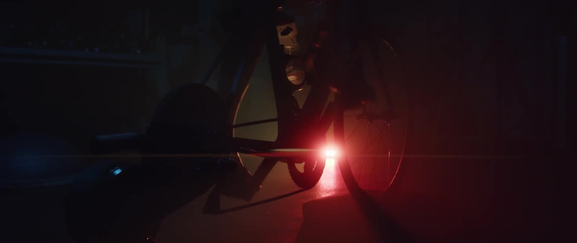 A person pedals on a stationary bicycle in a dark room illuminated by a colored light