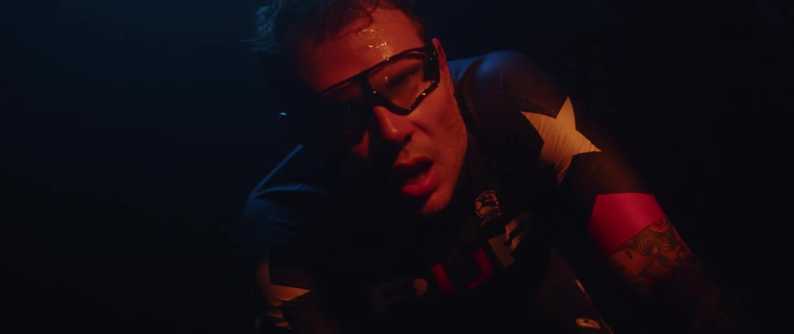 Man breathes and drops his head tiredly as he sits on stationary bike in a dark room