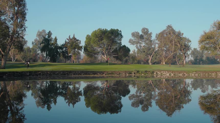 A person slowly walks while carrying golf clubs next to a reflective pond