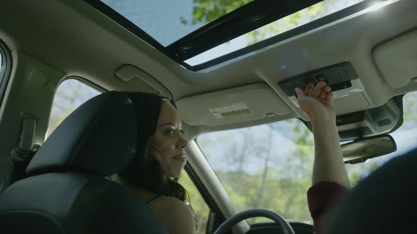 A teenage girl sitting in the driver seat of a car looks up as her passenger opens the sunroof