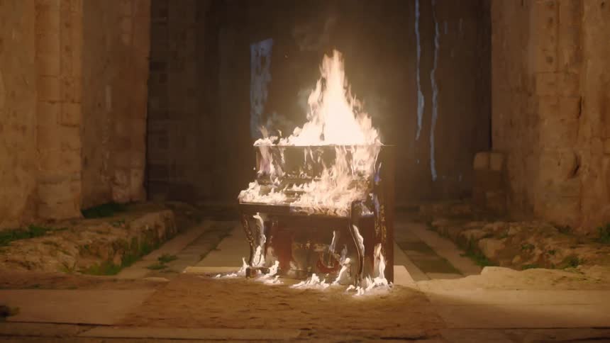An upright piano is engulfed in flames within a temple like building