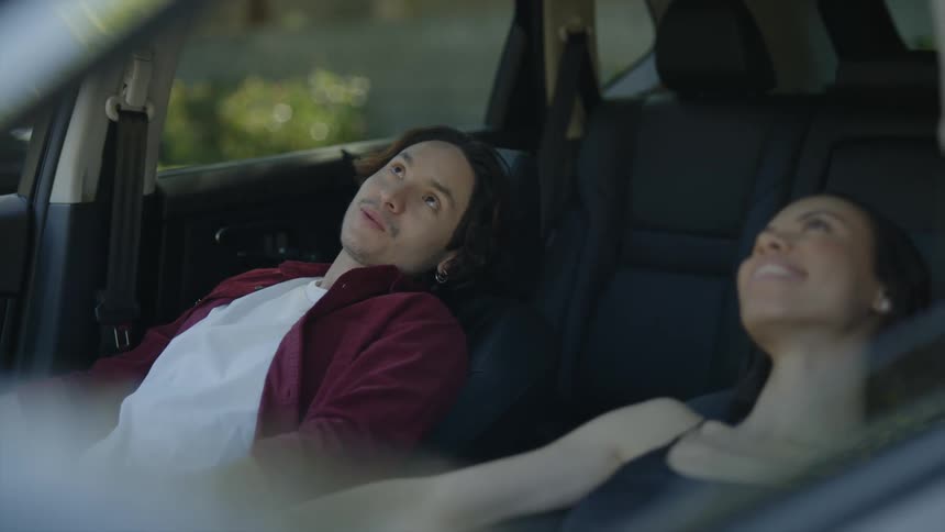 A couple leans back in their seats in a car and talk and look out the sun roof