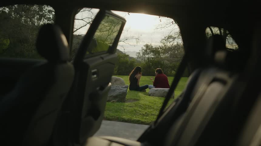 Young couple seen through a car sitting on grass talking overlooking the sunset
