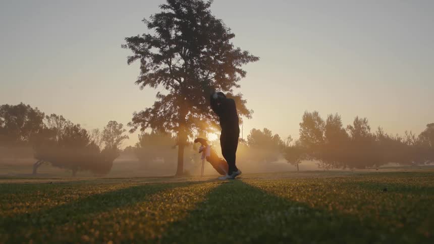 A man wearing a hat is standing in a field near a tree and hits a ball with his golf club as the sun shines brightly behind him in a hazy sky