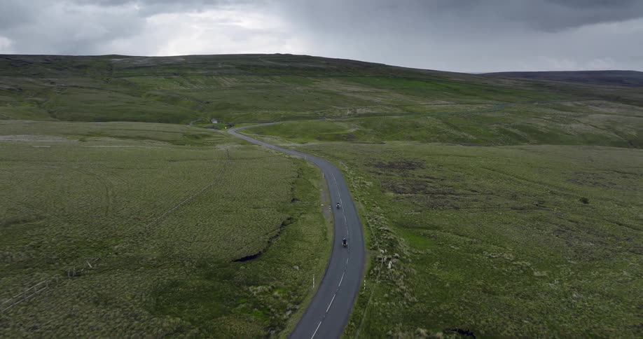 Motorcycles travel on a rural road in the country and there is a mountain in the distance under a cloudy sky