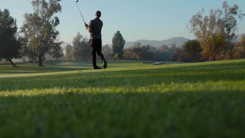Man standing over the green grass of a golf course hits a ball with a club at morning