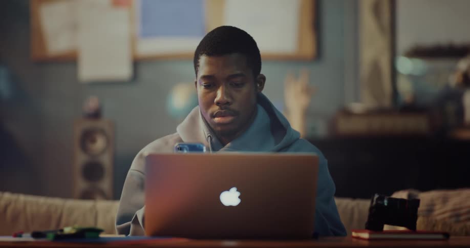 A man is sitting in front of a laptop computer holding a camera and he puts it down and picks up his cell phone