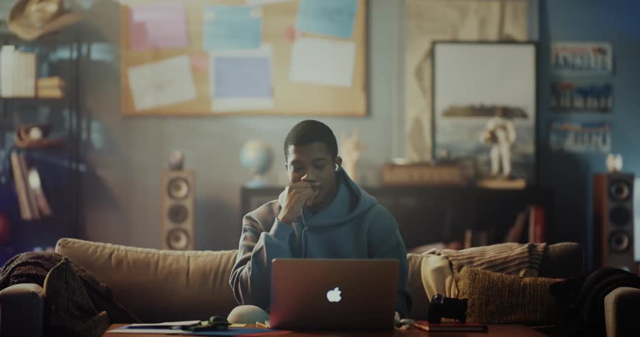A man sitting on a couch with his laptop open and ear buds in picks up a camera smiling and dancing