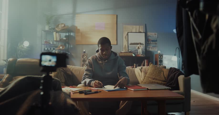 A man sits at home in front of a set up camera and writes on a piece of paper and then gets up and walks away