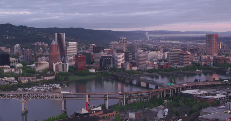 Traffic moves across a bridge as big city buildings and skyscrapers reflect on the water and there are mountains in the distance under a cloudy sky