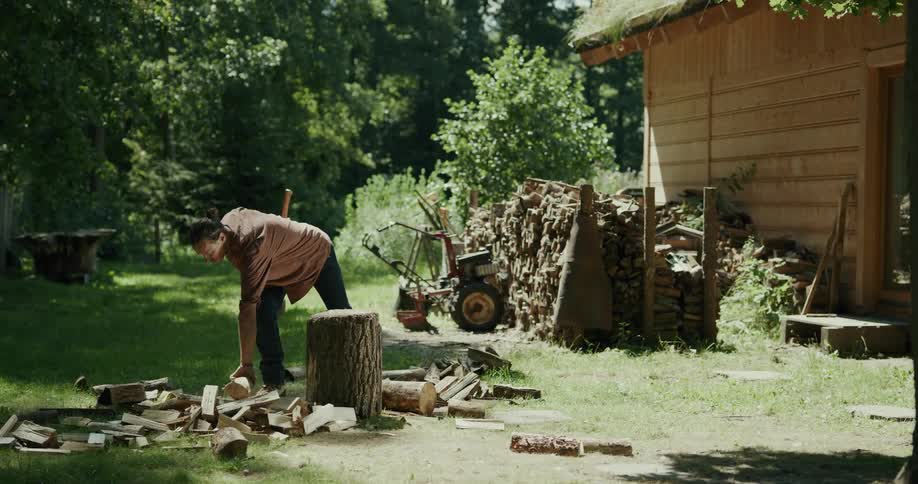 A tall man wielding an ax chops wood while outside of his house