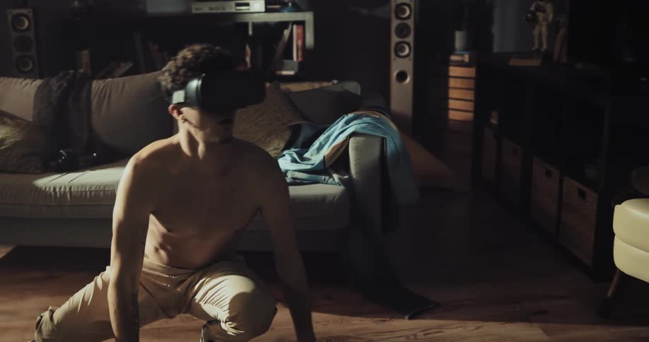 Young shirtless man crouches looking around in the living room while wearing vr goggles