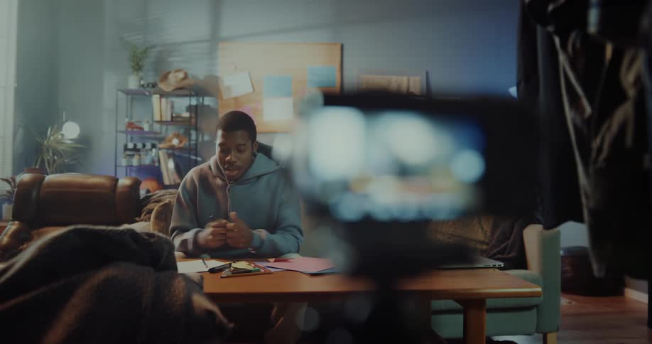 A man turns on a camera mounted on a tripod before sitting on a couch in front of it and beginning to speak