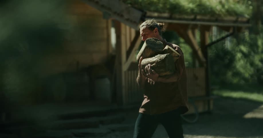 A man carries several logs near his cabin on a sunny day