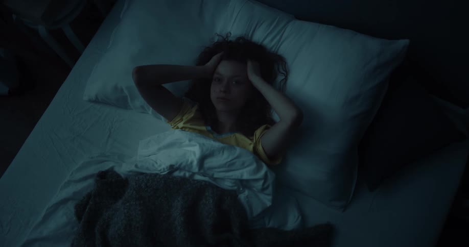 A woman is lying awake in bed holding her head and moving her fingers through her hair as lightning flashes