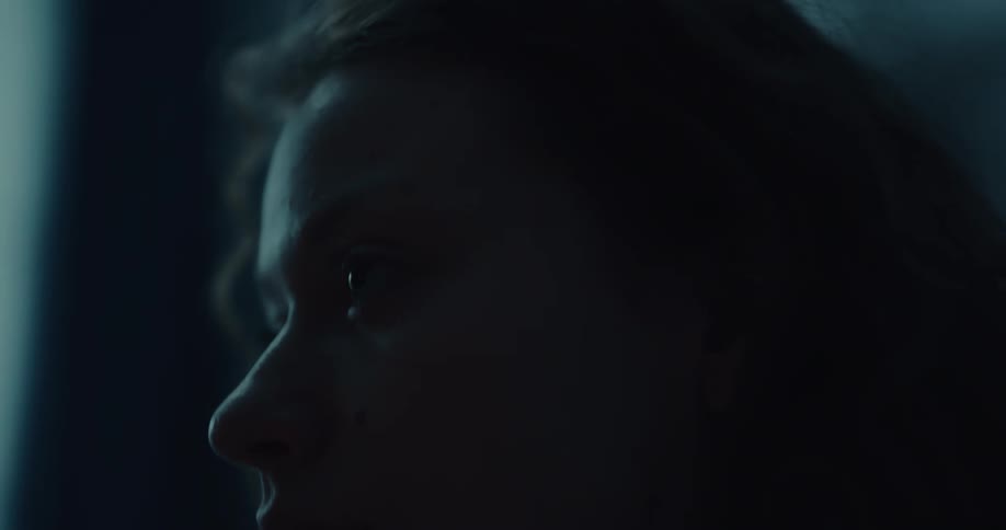 A contemplative young woman runs her hand through her hair while sitting in a dimly lit room