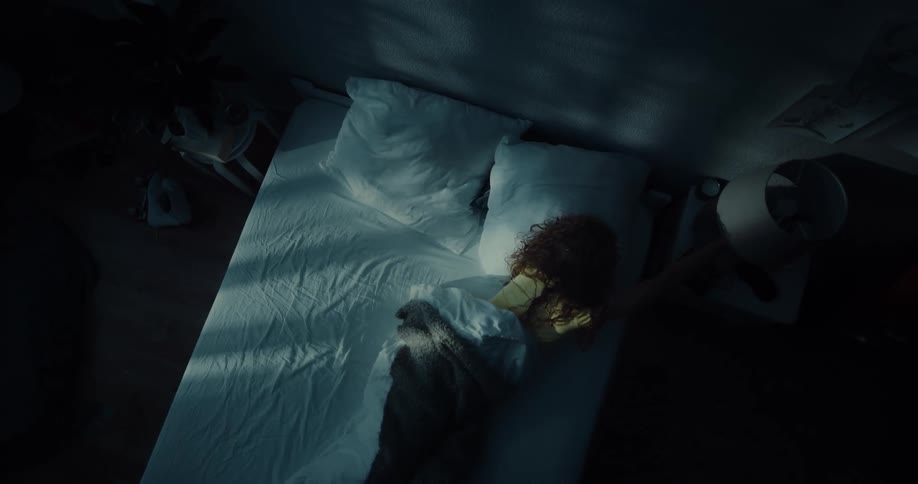 A young woman wakes up and turns on the lamp on her nightstand before lying back and gazing up at the ceiling
