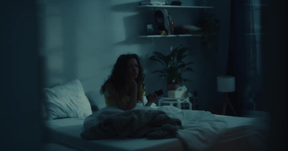 A young woman sits up in bed and uses a television remote control while looking bored