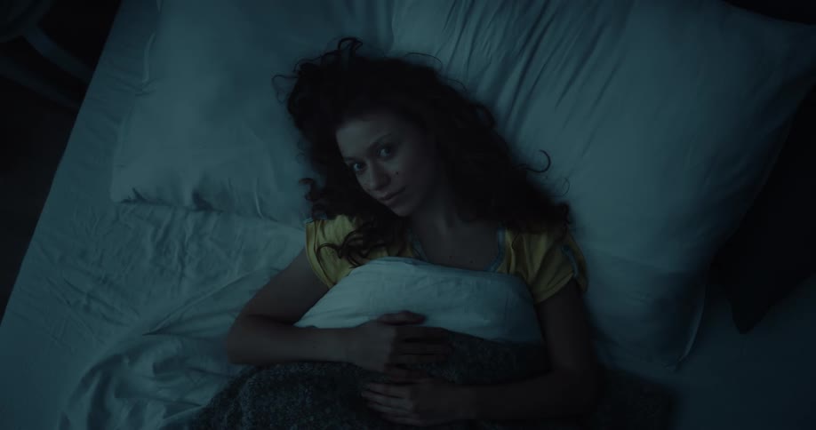 A woman raises her gaze to look into the camera while resting in bed in a dark bedroom