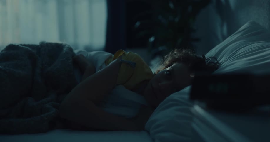 A woman rolls over in bed and there is an alarm clock on the nightstand next to her