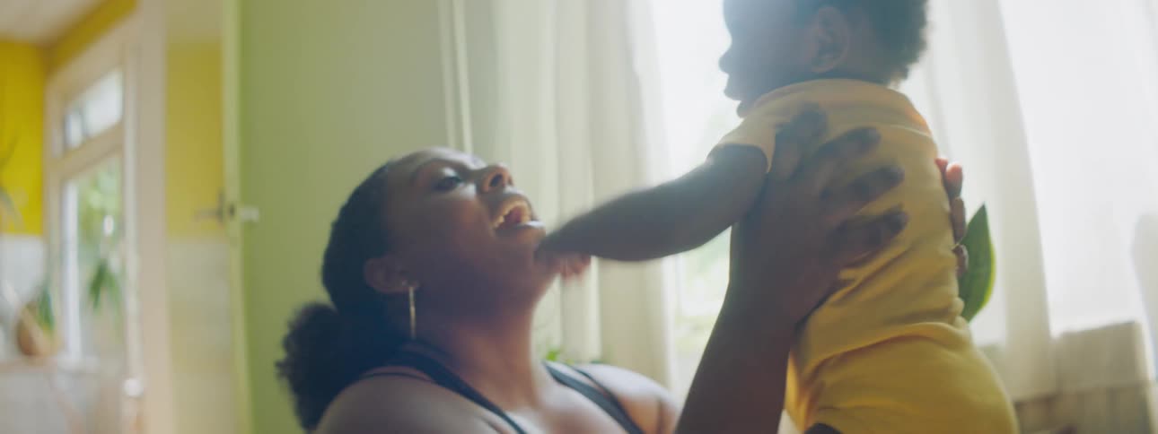 Mother with curly hair holds and kisses her toddler son wearing yellow clothes in the living room at home