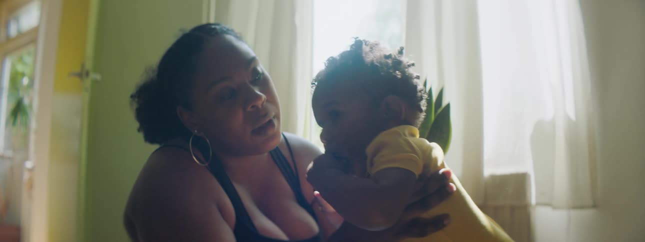 Mother with hoop earrings holds her toddler girl with yellow clothes and talks to her in the living room at home