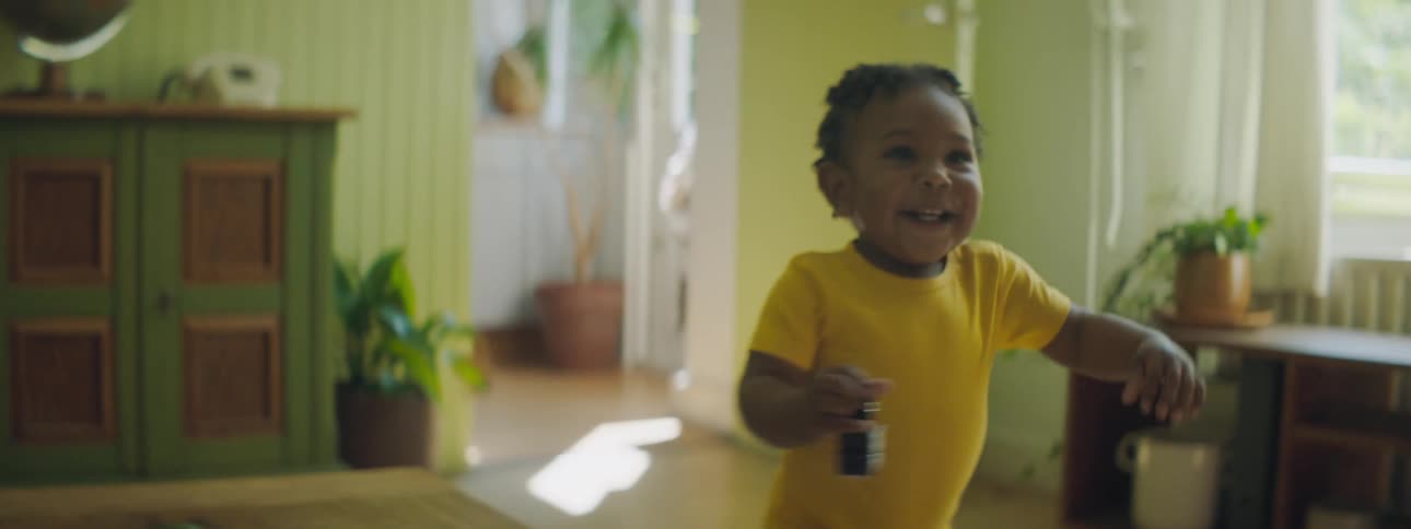 Toddler in yellow onesie walks across wooden floor past window