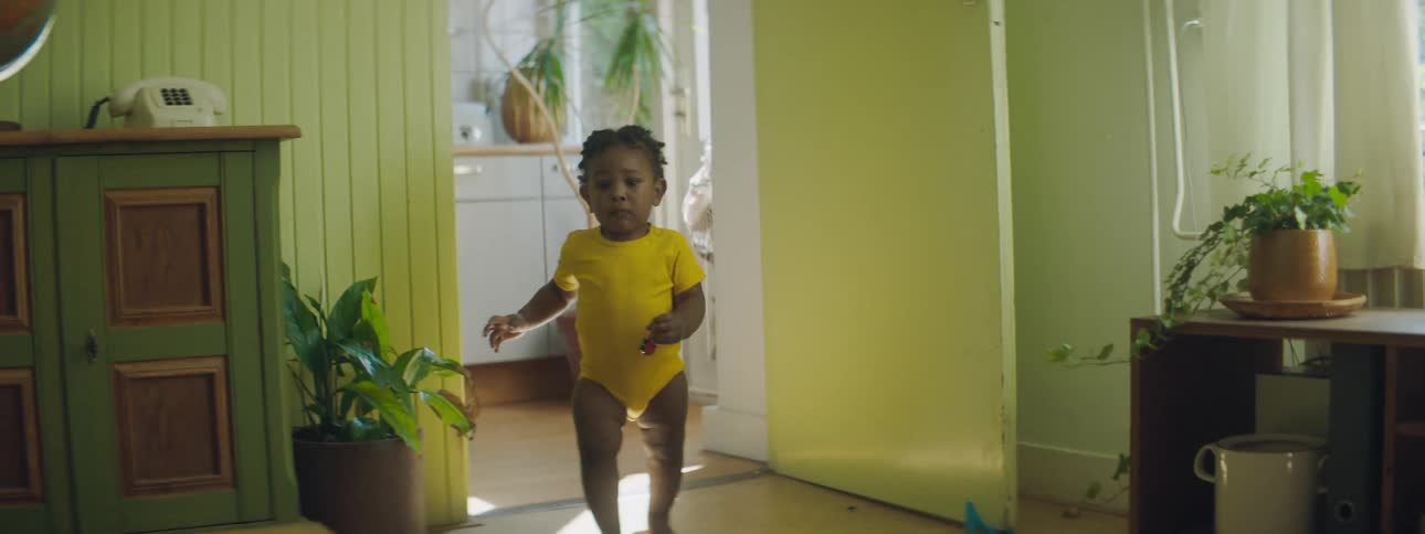 Toddler wearing yellow onesie walks from kitchen into room past cabinet