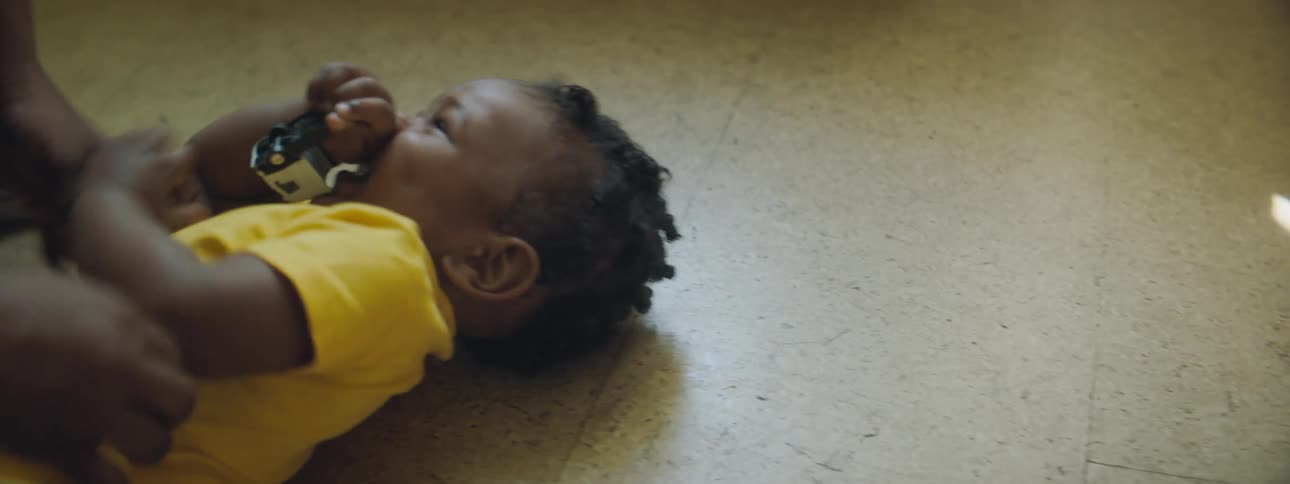 A happy african american baby wearing a yellow onesie is laying on the floor being tickled by a older person