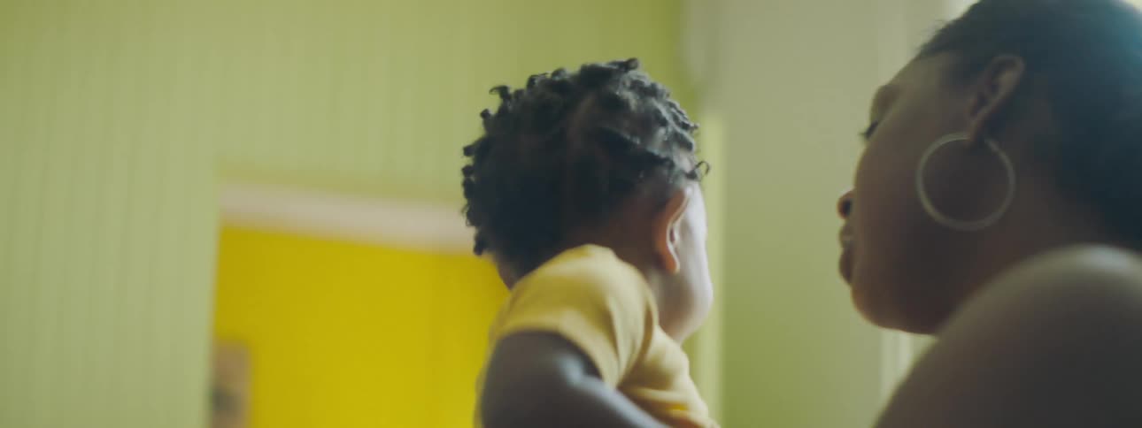Mother with and undershirt and hoop earrings holds and kisses her toddler girl with yellow clothes