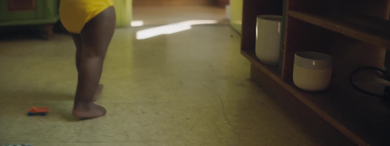 A young baby walks across the living room helped by an adult holding their hands up