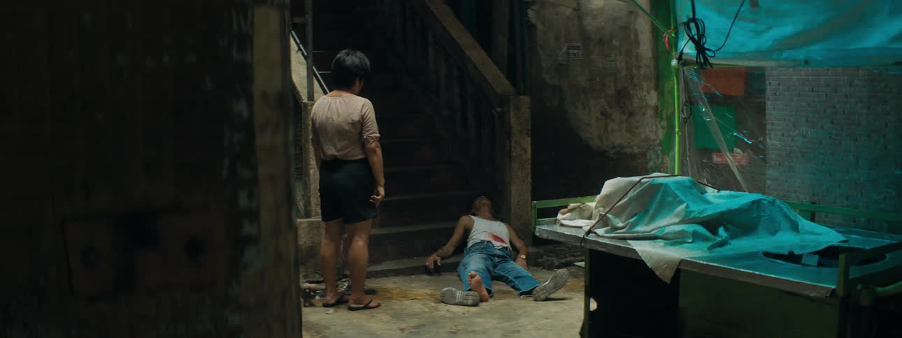 A woman stands and looks at a man lying at the bottom of a set of stairs not moving with blood on his shirt